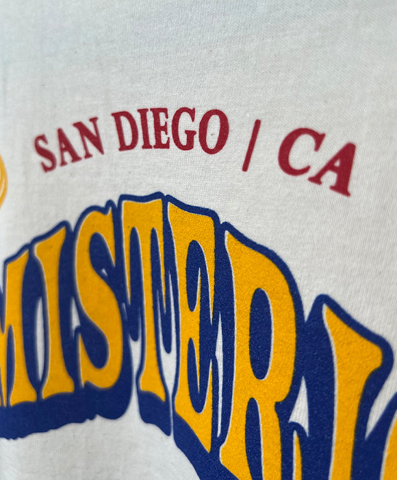 Text on a cream t-shirt with 'San Diego / CA' in red and 'El Misterio de Chula Vista' in yellow and blue.
